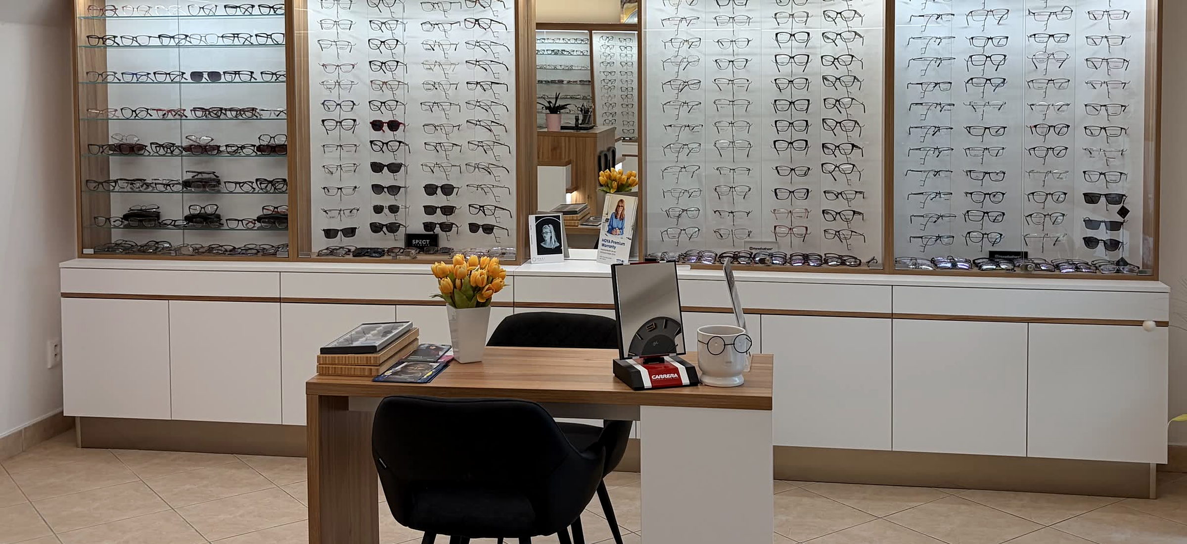 Konzultační koutek prodejny Optik Dvořák — dřevěný stůl se dvěma černými židlemi, stolním zrcadlem a vázou se žlutými tulipány, kolem dokola stěny s regály plnými brýlových obrub